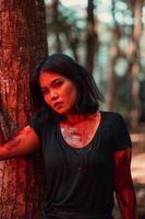 an Asian woman in black clothes and covered in blood stood in front of a tree with a very scary expression photo