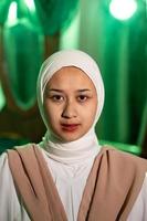 a Muslim woman with a white headscarf and white clothes is sitting in the middle of a green room without makeup photo