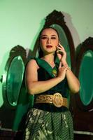 a female Javanese dancer in a green dance costume and a full face with makeup poses with her hands in front of a dressing table photo