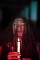 an Asian woman with closed eyes and a transparent veil stained with blood holds a candle in her hand during satanic worship photo