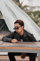 an Asian man with a chubby face wearing sunglasses and a black leather jacket while sitting at a cafe table photo