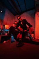 a couple of gay teenagers are sitting together wearing sunglasses and posing naughty in a warehouse with a red light photo