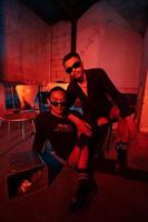 a couple of gay teenagers are sitting together wearing sunglasses and posing naughty in a warehouse with a red light photo