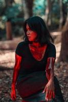 un hembra de serie asesino en negro ropa y cubierto en sangre tiene un de miedo cara sentado en el medio de un bosque foto