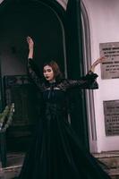 an Asian woman with a witch-like appearance flung her all-black skirt in the air as she stood in front of a cemetery photo