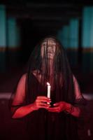 un asiático mujer con cerrado ojos y un transparente velo manchado con sangre sostiene un vela en su mano durante satánico Adoración foto