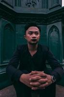 an Asian man in a black suit is sitting alone in front of the funeral monument with a very scary expression photo