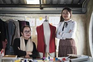 Moda equipo gente, asiático hembra diseñador y adolescente asistente en estudio, brazos cruzado y sonrisa, contento trabajando con hilo y de coser para vestir diseño, profesional boutique Sastre SME emprendedor.