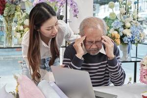 Asian elderly male florist owner is unwell with headaches and health deteriorating, stress from overwork, young female worker support with care in a flower shop, family SME business entrepreneur. photo