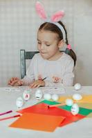 un pequeño niña en conejito orejas en su cabeza decora blanco huevos con pegatinas con diferente emociones, en su manos es un blanco huevo con emoción foto