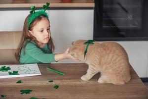 A little girl with a rim decorated with clover draws and cuts green shamrocks for St. Patrick's Day at the table at home in the kitchen, next to her is her beautiful cat photo