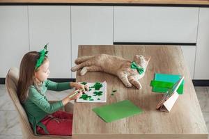 A little girl with a bandage on her head draws and cuts green shamrocks for St. Patrick's Day at a table at home in the kitchen, next to her is her beautiful cat with a green bow tie around his neck photo