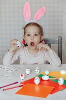 a little girl in bunny ears on her head decorates white eggs with stickers with different emotions, in her hands is a white egg with emotion, the child copies and shows with facial expressions. photo