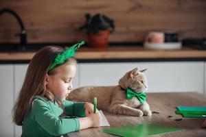 A little girl with a bandage on her head draws and cuts green shamrocks for St. Patrick's Day at a table at home in the kitchen, next to her is her beautiful cat with a green bow tie around his neck photo