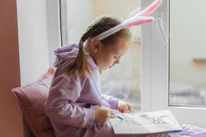 una colegiala con orejas de conejo se sienta en la ventana y dibuja un conejo en su álbum, preparándose para pascua foto