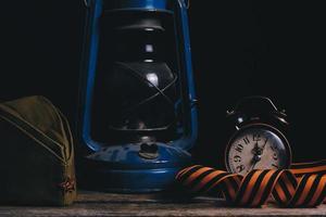 Russian soldiers cap, St. George ribbon, an old retro alarm clock and kerosene lamp dark room on wooden table.