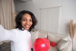 selfie retrato de feliz joven afroamericana con globo de corazón rojo. día de san valentín 14 de febrero o cumpleaños en apartamento moderno foto