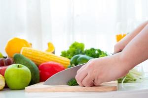 Manos usando un cuchillo para picar verduras sobre tabla de talla de madera sobre la mesa en el fondo de la cortina blanca foto