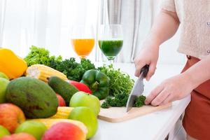 Manos usando un cuchillo para picar verduras sobre tabla de talla de madera sobre la mesa en el fondo de la cortina blanca foto