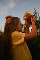 A happy young girl with long hair in a straw hat stands in a large field of sunflowers. Summer day. A warm sunset. photo