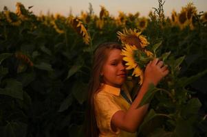 A happy young girl with long hair in a straw hat stands in a large field of sunflowers. Summer day. A warm sunset photo