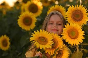 un contento joven niña con largo pelo en un Paja sombrero soportes en un grande campo de girasoles verano día. un calentar puesta de sol. foto