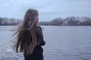 un niña con largo pelo en el apuntalar de un lago. en pie en el viento y mirando a un hermosa calentar puesta de sol. foto