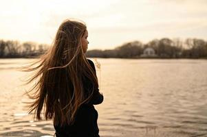 un niña con largo pelo en el apuntalar de un lago. en pie en el viento y mirando a un hermosa calentar puesta de sol. foto