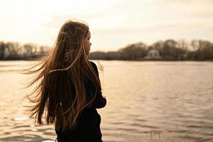 A girl with long hair on the shore of a lake. Standing in the wind and looking at a beautiful warm sunset. photo