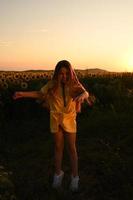 un contento joven niña con largo pelo en un Paja sombrero soportes en un grande campo de girasoles verano día. un calentar puesta de sol foto