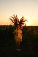 un contento joven niña con largo pelo en un Paja sombrero soportes en un grande campo de girasoles verano día. un calentar puesta de sol. foto