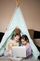 Two girls sisters watching on laptop at wigwam tent. Technology and home concept. photo