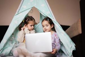 dos muchachas hermanas acecho en ordenador portátil a wigwam carpa. tecnología y hogar concepto. foto