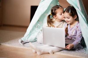 dos muchachas hermanas acecho en ordenador portátil a wigwam carpa. tecnología y hogar concepto. foto