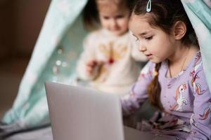 Two girls sisters watching on laptop at wigwam tent. Technology and home concept. photo