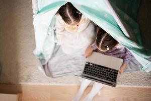 dos muchachas hermanas acecho en ordenador portátil a wigwam carpa. tecnología y hogar concepto. foto