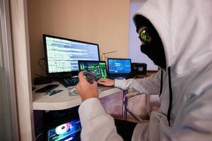 Boy hacker fraudulently use credit card for payment. Internet theft . Man wearing a balaclava and holding a credit card while sat behind a laptop. photo