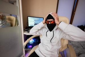 Boy hacker grabbed his head. Internet theft . Man wearing a balaclava sat behind a laptop. photo