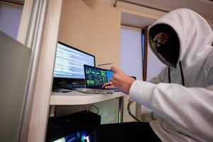 Boy hacker fraudulently use credit card for payment. Internet theft . Man wearing a balaclava and holding a credit card while sat behind a laptop. photo