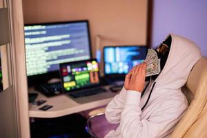 Boy hacker sniffs money dollars. Internet theft . Man wearing a balaclava sat behind a laptop. photo
