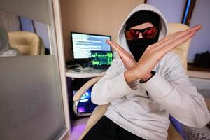 Boy hacker fraudulently use credit card for payment. Internet theft shows cross hands . Man wearing a balaclava and holding a credit card while sat behind a laptop. photo