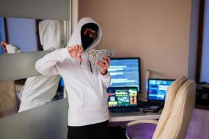 chico hacker con dinero dólares, espectáculo abajo dedo. Internet robo . hombre vistiendo un pasamontañas detrás un ordenador portátil. foto
