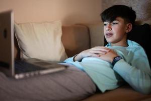 joven adolescente chico en frente de un ordenador portátil en un cama a noche. foto