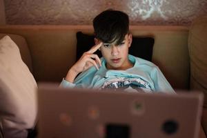 Young teen boy in front of a laptop on a bed at evening. photo