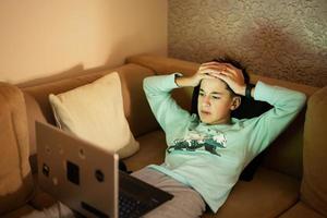 Young teen boy in front of a laptop on a bed at evening watching football match. photo