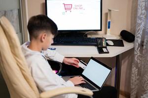 Happy teenage boy using laptop and credit card, sitting at desk and ordering goods online from home. photo