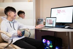 contento Adolescente chico utilizando ordenador portátil y crédito tarjeta, sentado a escritorio y ordenando bienes en línea desde hogar. foto