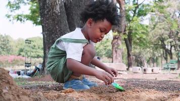 un africano americano niño cavar un agujero en el arena con un juguete espátula en patio de recreo en parque video
