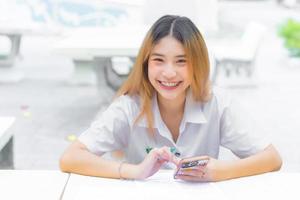 joven estudiante asiática en uniforme usando un teléfono inteligente y escribiendo algo sobre el trabajo. Hay muchos documentos sobre la mesa, su rostro sonriendo en un trabajo para buscar información. foto