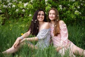 dos joven muchachas en vestidos son sentado en el verde césped debajo un blanco árbol y riendo foto